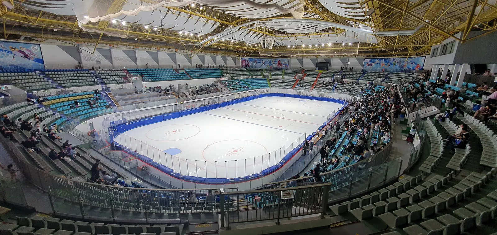 The empty ice of Mokdong Ice Rink seen from the stand, with the blue boards, the faceoff markings and the spectators' seats around it (June 2023). — Mokdong Ice Rink