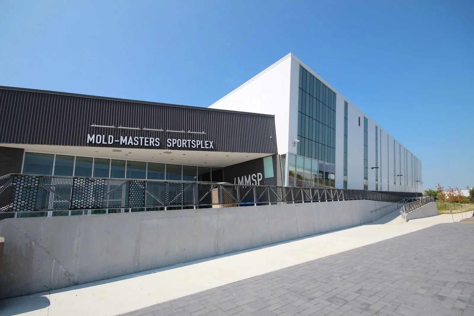 The Mold-Masters SportsPlex entrance, its name across a dark canopy beside a tall white rink block with a column of glazing, a ramp in front. — Mold-Masters SportsPlex