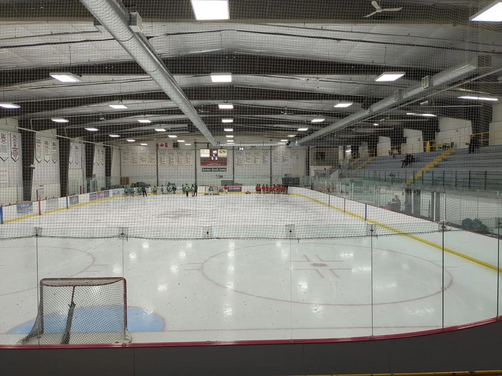 A Mold-Masters SportsPlex ice pad from behind the net and glass, with a few skaters and the stands along the far side. — Mold-Masters SportsPlex