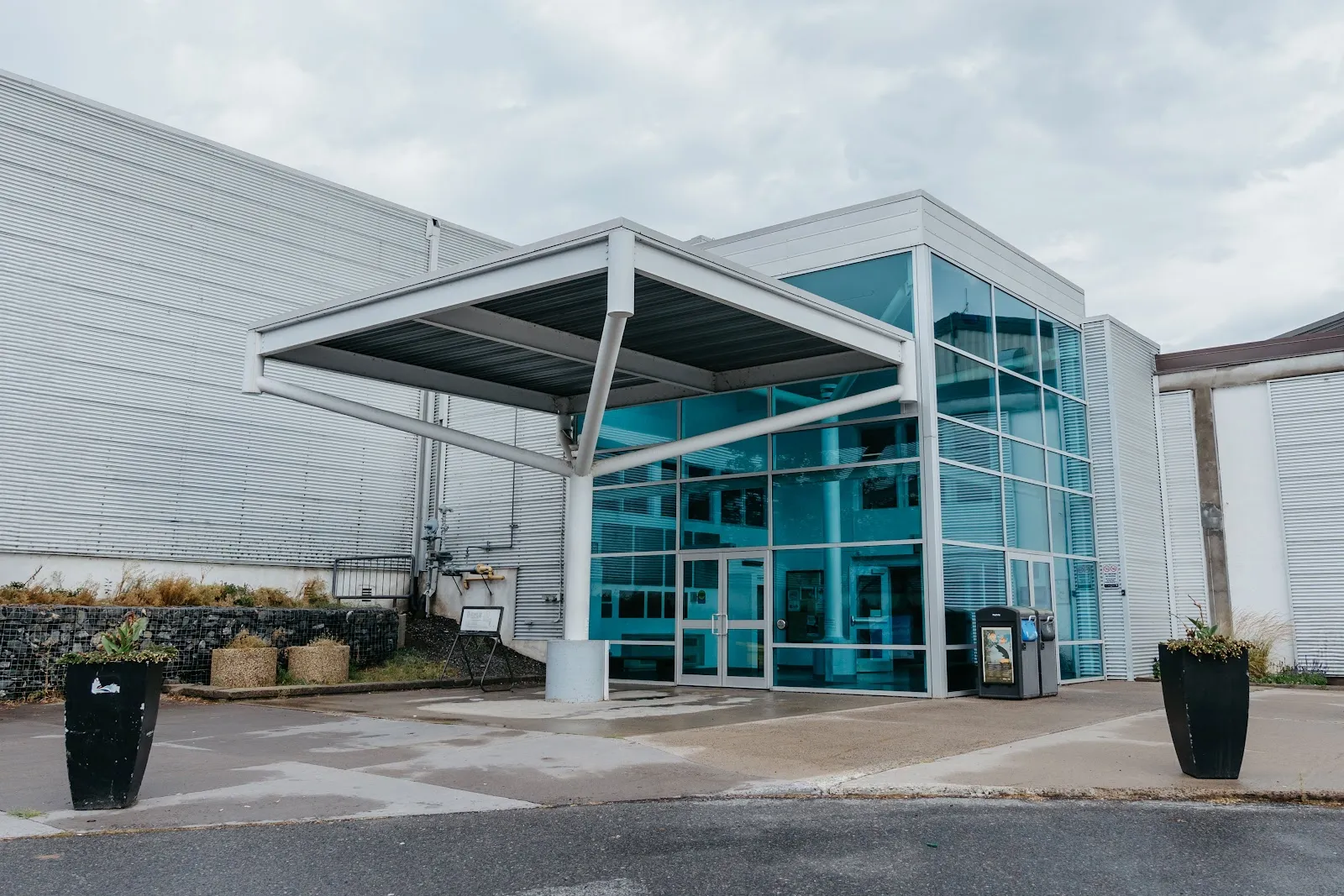 The glass-fronted entrance of the Moncrief Construction Sports Centre with a flat steel canopy, tinted blue-green windows and black planters, set against the building's white corrugated wall under a cloudy sky. — Moncrief Construction Sports Centre (Kenora)