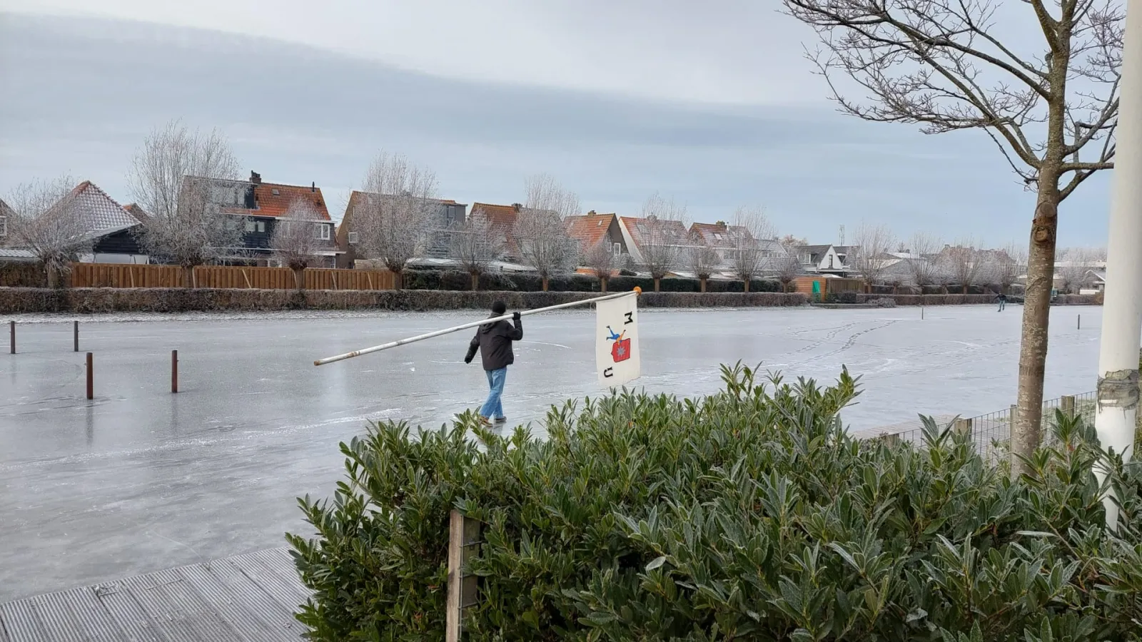 The natural-ice rink at Moordrecht on opening day, a skater in the distance and a man with his back to the camera carrying the club flag, with rows of houses and pollard willows along the far bank. — IJsbaan Moordrecht (Moordrechtse IJsclub)