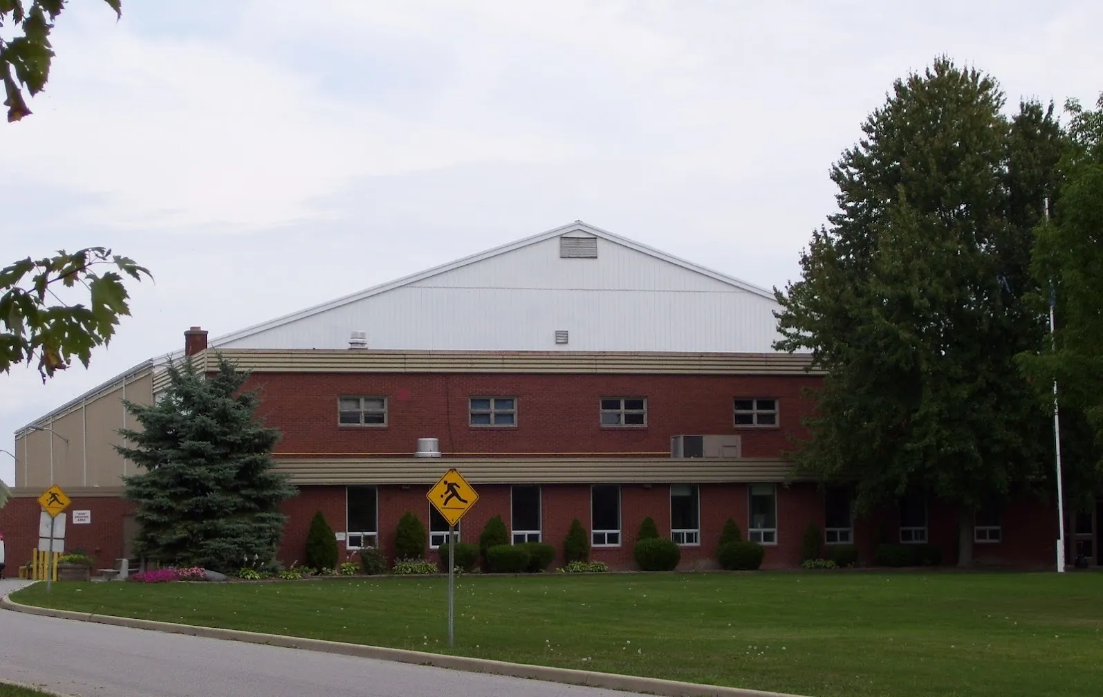 The two-storey red-brick front of the complex, with the arena's pale gabled roof rising behind it across a lawn. — Mooretown Sports Complex