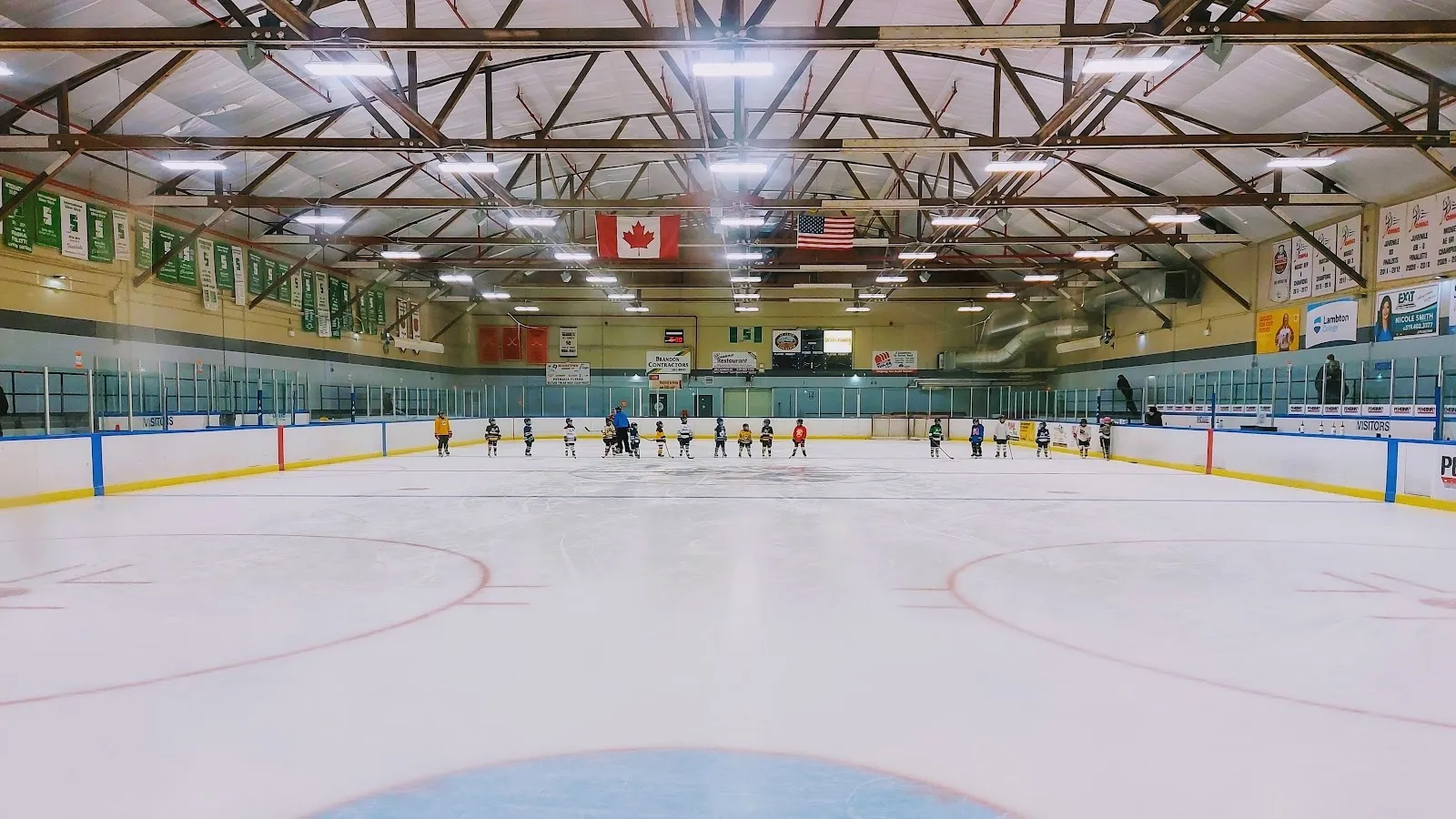 A wide view of the empty sheet from behind the glass, white ice with a blue crease and a long timber-trussed roof with a flag on the far wall. — Mooretown Sports Complex