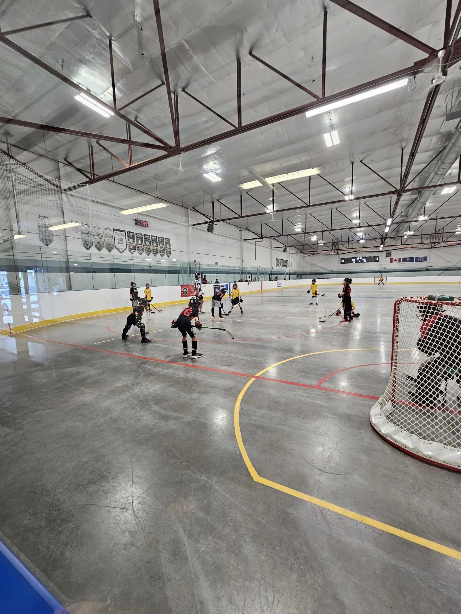 A game under a dark steel-trussed roof seen from the rail, players spread across a wide sheet with a net and yellow arc at the right. — Moose Recreation Centre