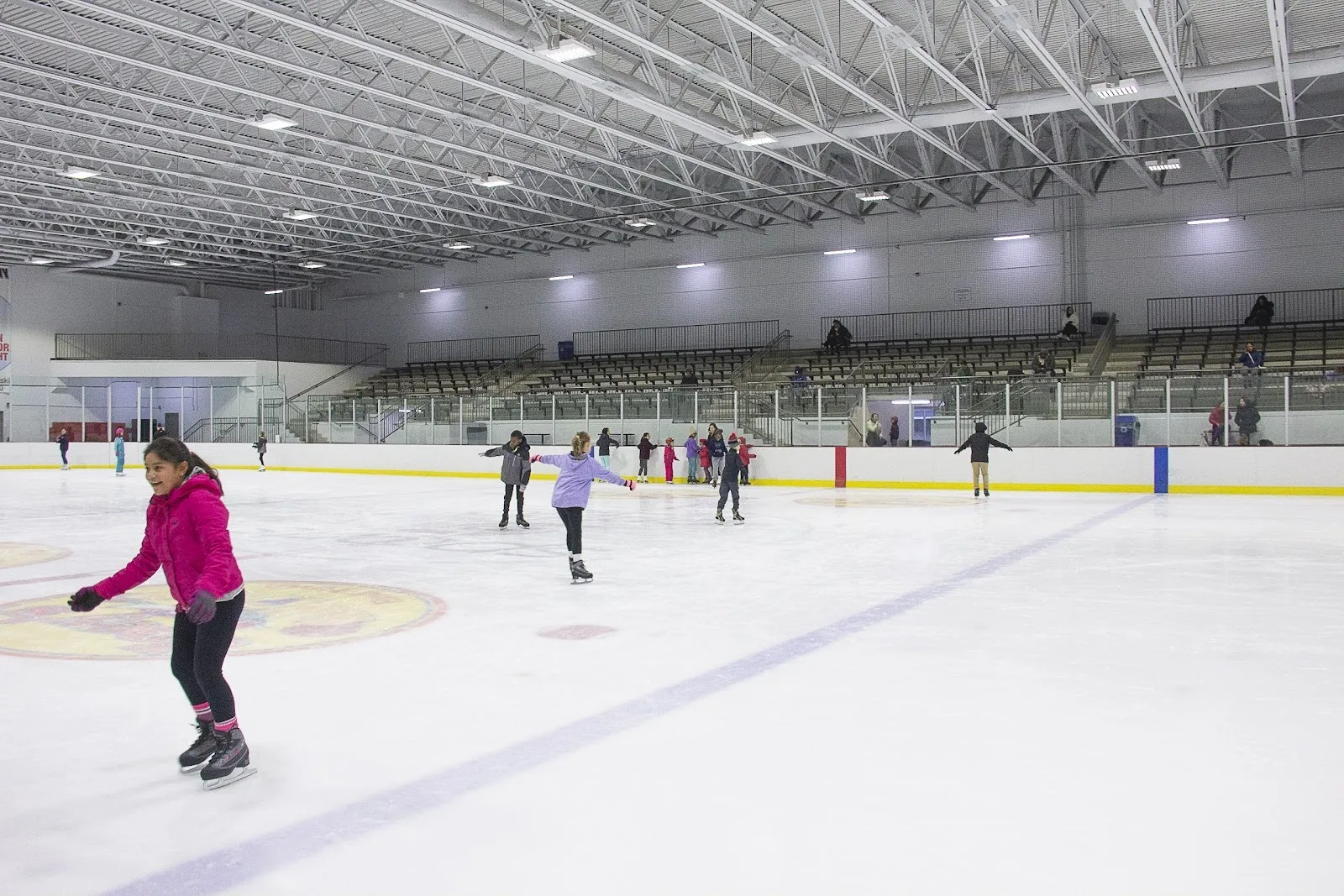 Una sesión pública en una pista amplia, con unos cuantos patinadores repartidos por el hielo bajo un largo techo de armaduras de acero, con gradas en el lado opuesto. — Morgan Park Sports Center (Chicago)
