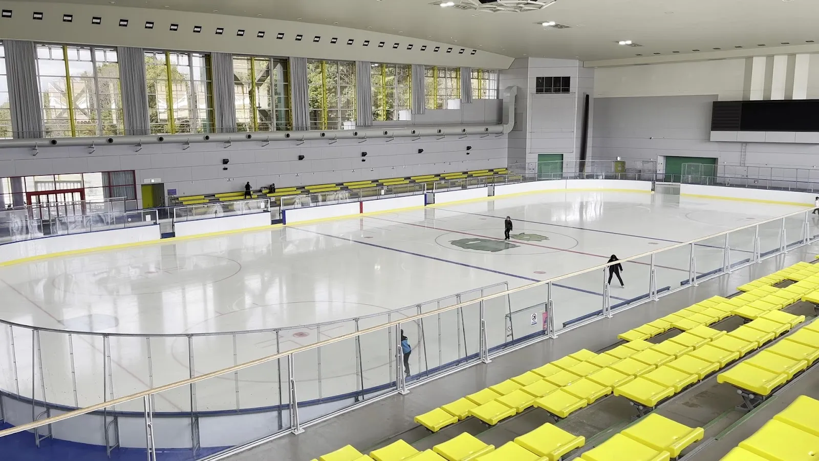 黄色い客席から見た、モリコロパーク アイススケート場の氷面。窓のある高い天井のホールで、氷の遠くに数人の滑走者がいます — Moricoro Park Ice Skating Rink