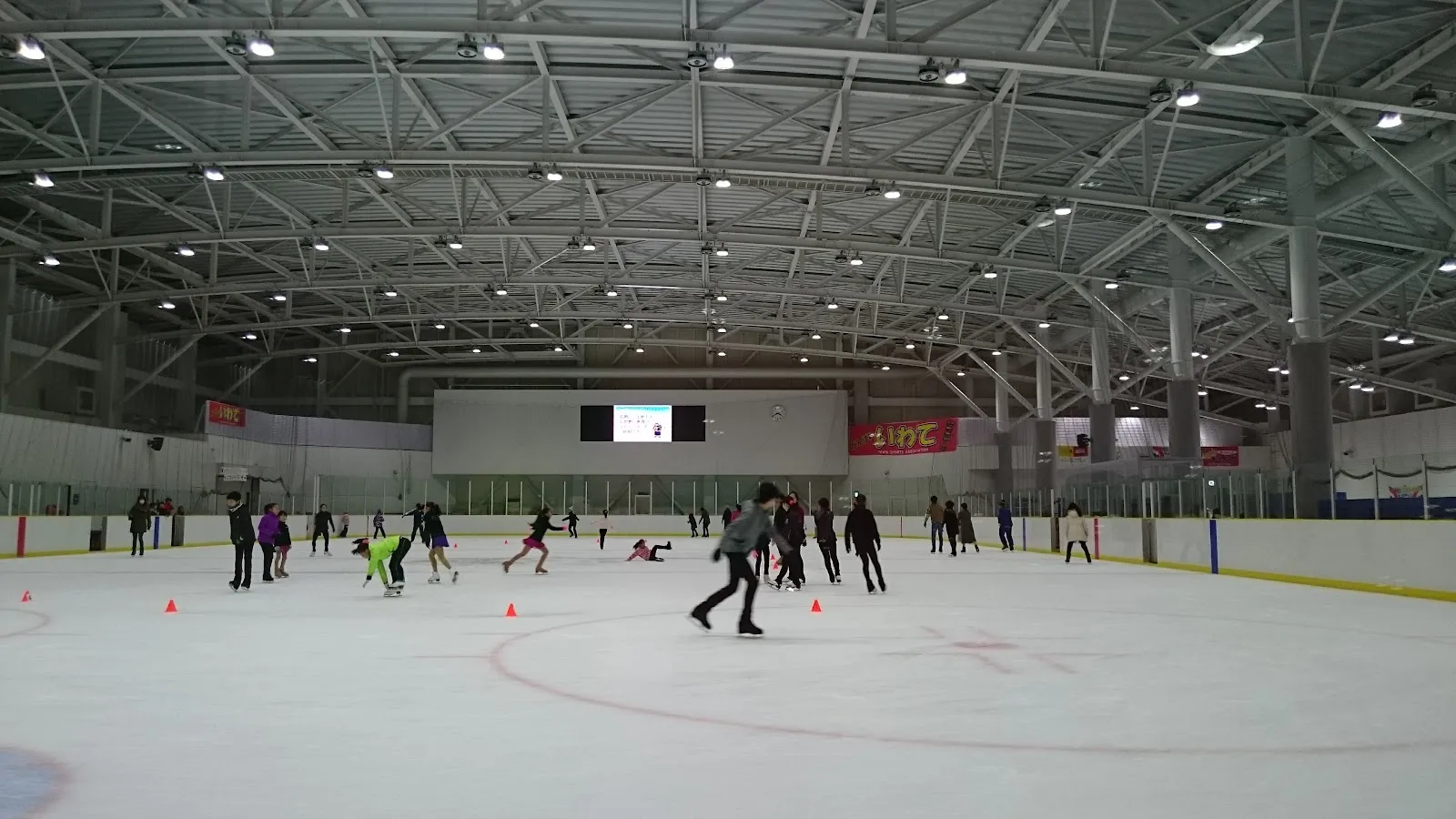 The main ice sheet of the Morioka City Ice Rink under its steel-trussed roof, with a few skaters weaving between training cones. — Morioka City Ice Rink