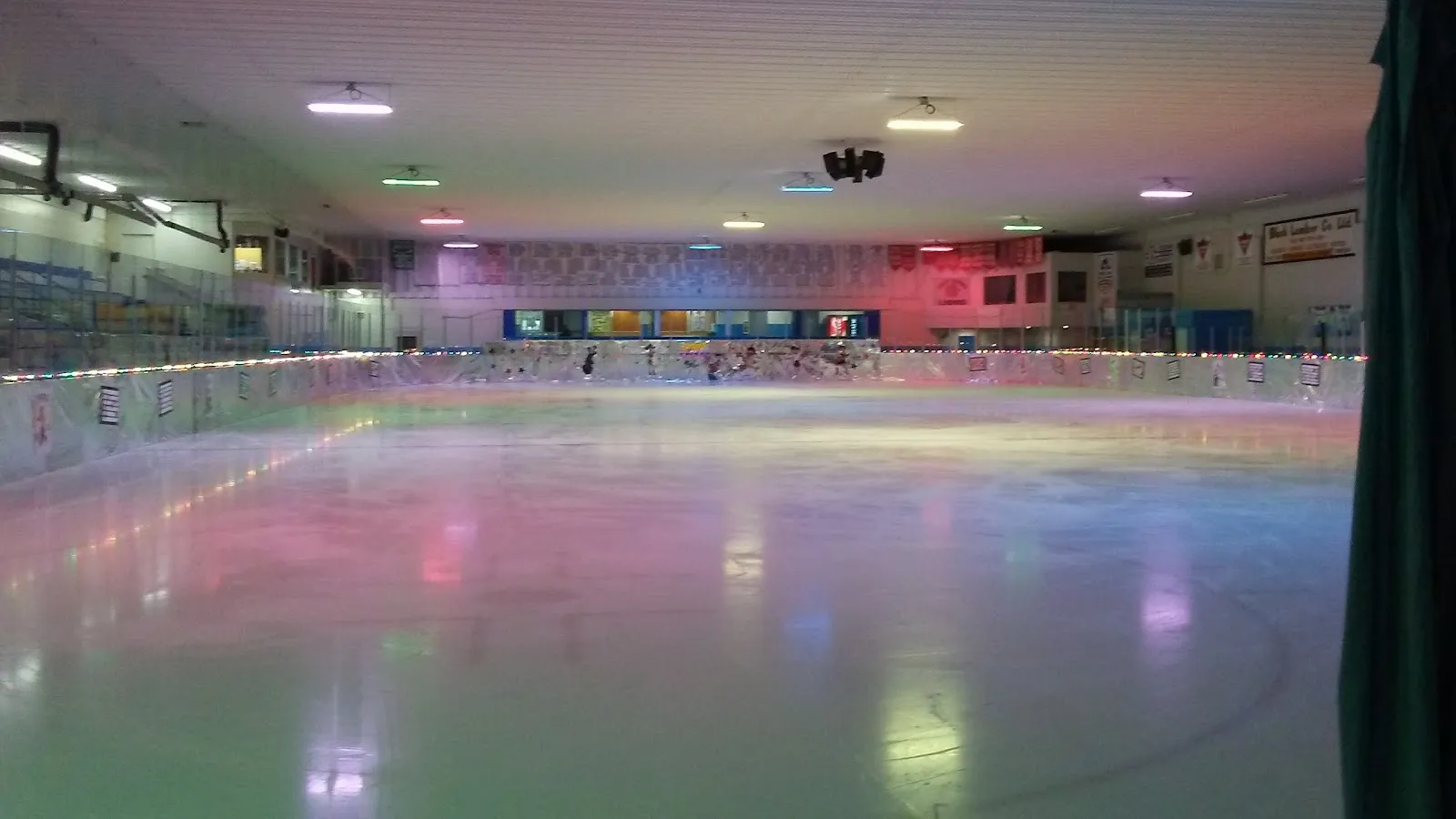 The empty ice sheet at the Morrisburg Arena lit by coloured lights, with the boards and the viewing windows along the far side. — Morrisburg Arena