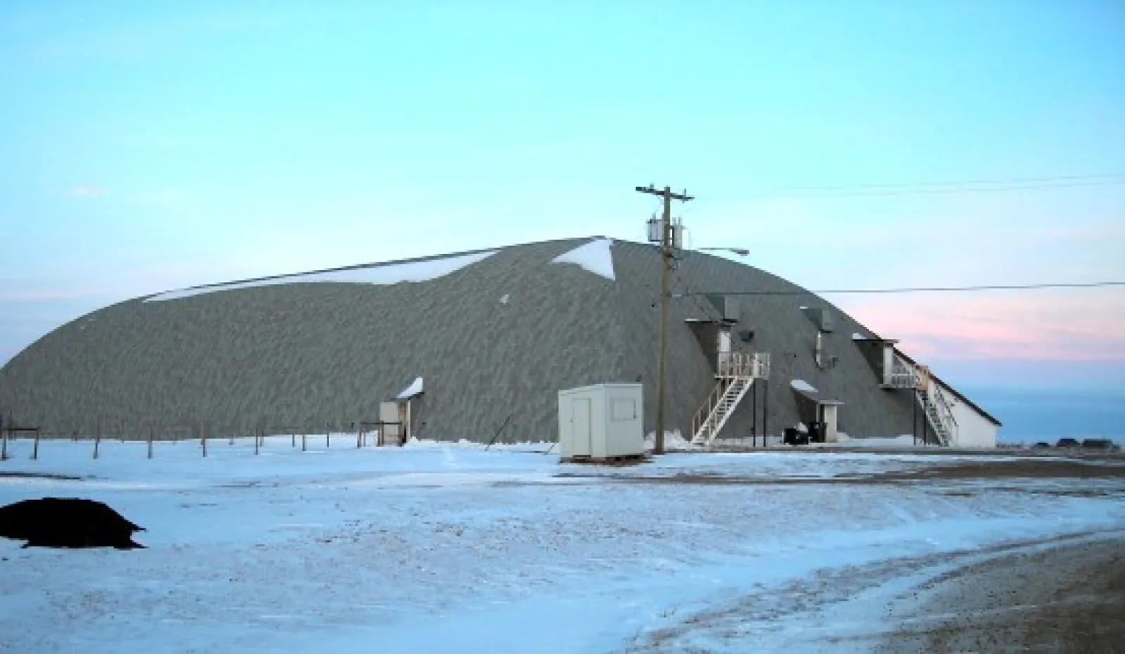 The Mortlach Centennial Rink building, a long arched metal-clad hall with outside stairs and a utility pole, seen across the snow at dusk. — Mortlach Centennial Rink