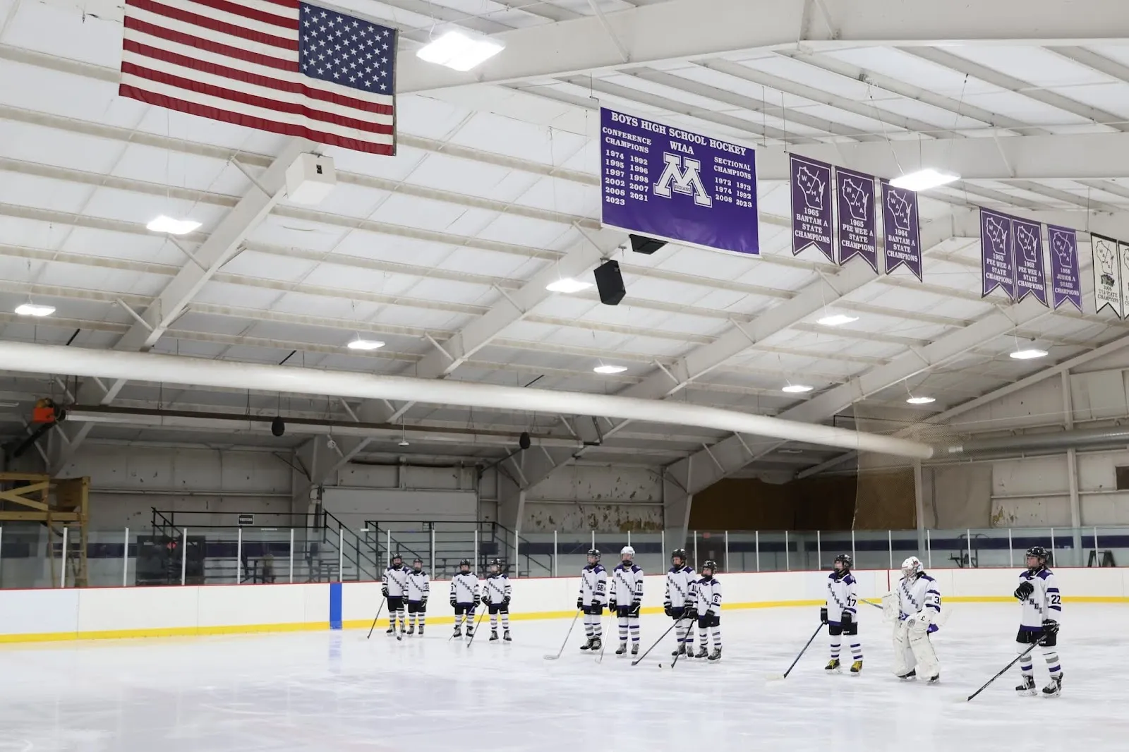 A team lined up along the blue line far down the sheet beneath an American flag and purple banners hung from a steel-trussed roof. — Mosinee Recreation Center