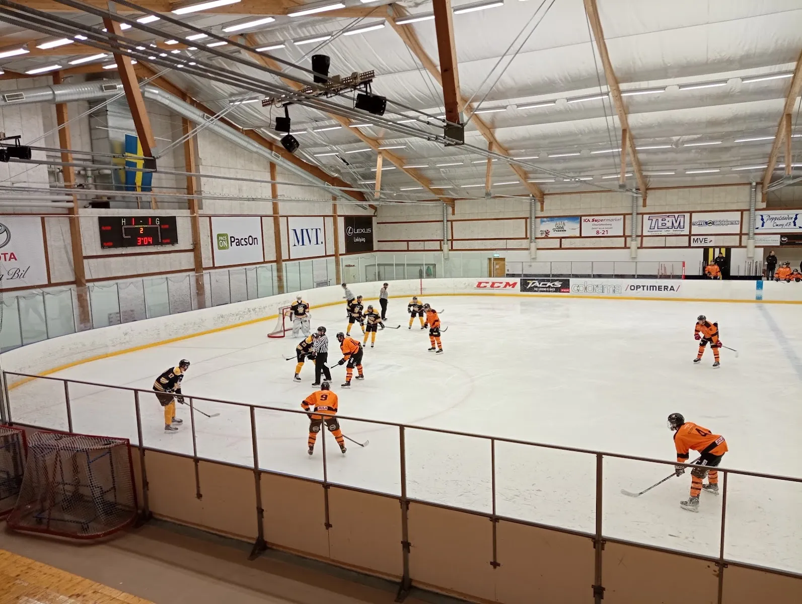 Ice hockey game in the hall at Motala ice stadium, players around the goal and the scoreboard on the far wall under the timber roof – Motala isstadion (Hargassner-hallen)