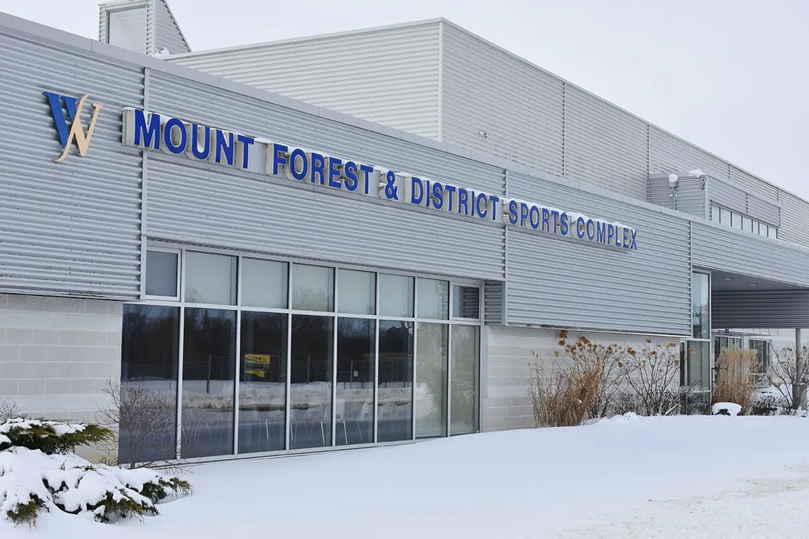 The grey-clad front of the Mount Forest & District Sports Complex in winter, its name in blue letters above the glass lobby. — Mount Forest & District Sports Complex