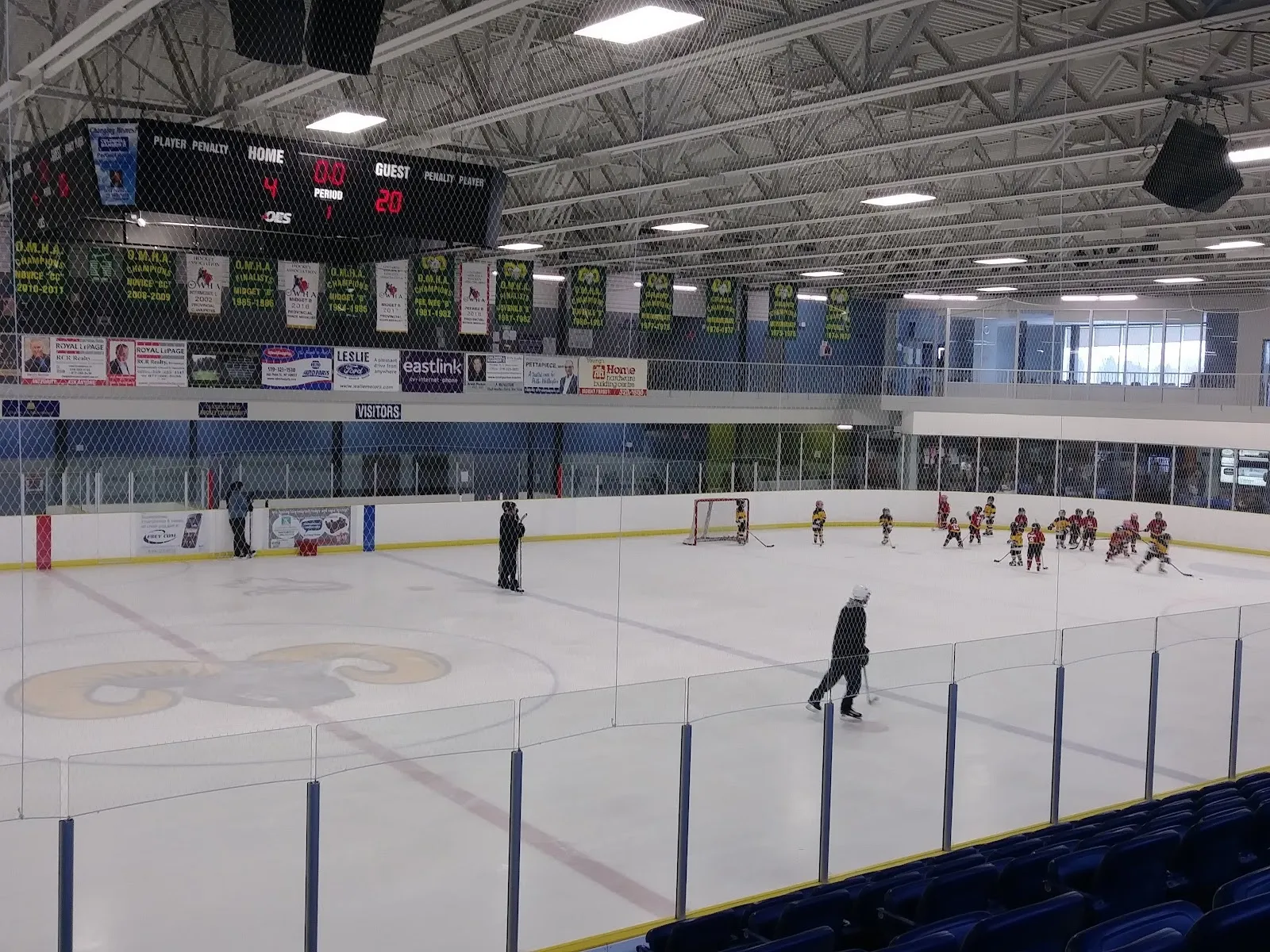 A game seen from the blue seats, players spread across the sheet under a steel roof and banners along the far wall. — Mount Forest & District Sports Complex
