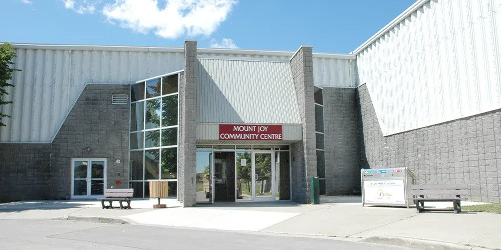 Front entrance of the building, a metal-clad structure with grey block piers, glass entry doors, and a red sign reading 'MOUNT JOY COMMUNITY CENTRE' above the doors. — Mount Joy Community Centre