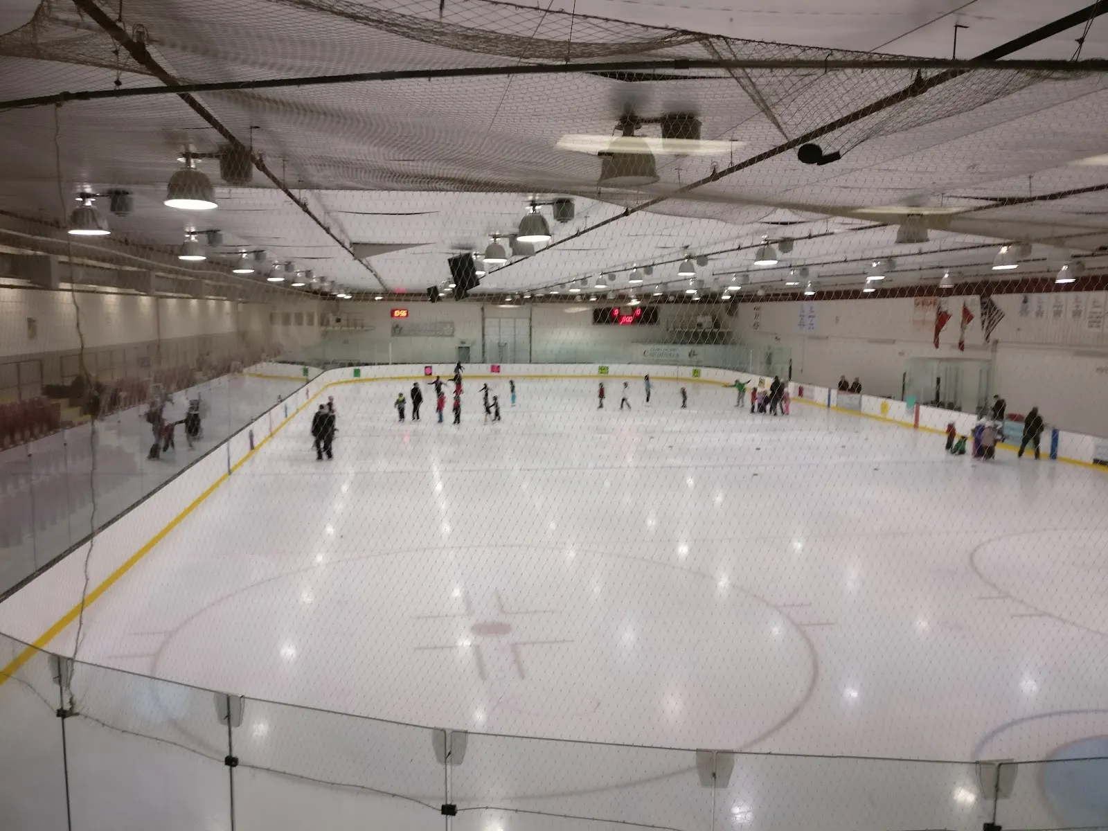 A wide view from the stands of the sheet with red lines and a faceoff circle, players spread near the far boards under a long steel roof. — Mount Joy Community Centre