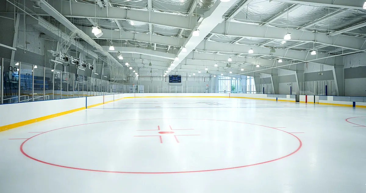 An empty NHL-size sheet in the rebuilt arena, seen from center ice, with glass boards, a scoreboard and a high steel-truss ceiling. — Mount Vernon Rec Center Ice Arena