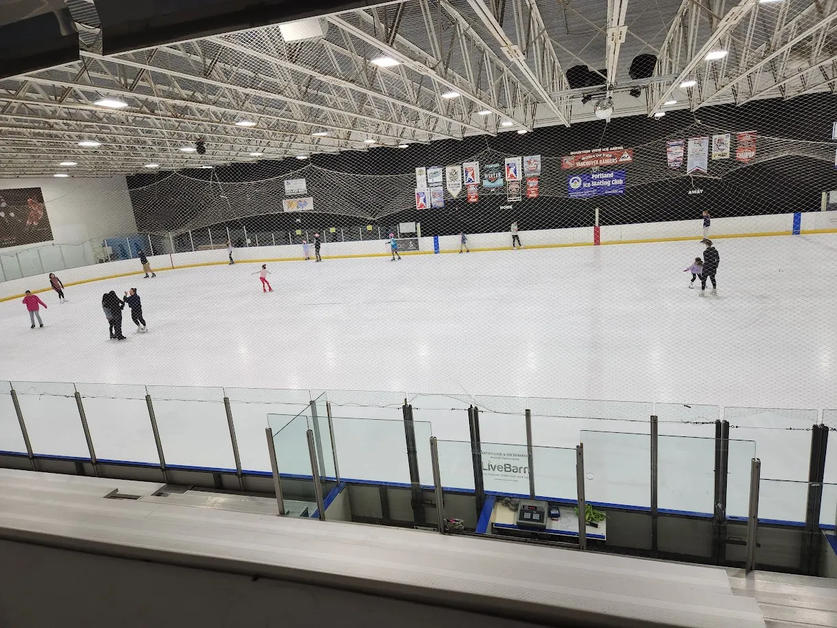 Elevated view from the stands of the full ice sheet during a public session, with MOUNTAIN VIEW ICE ARENA, Vancouver Rangers, and Portland Ice Skating Club banners hanging above the black padded boards and several skaters visible in the distance. — Mountain View Ice Arena