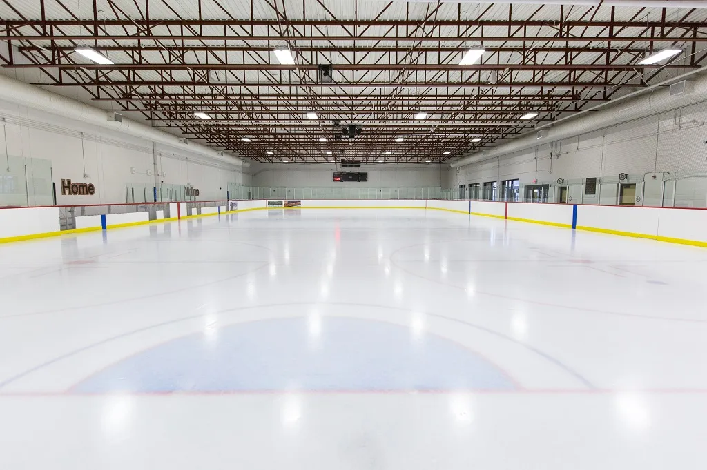 Full ice sheet at Mountainside Arena viewed from the boards, empty rink with "Home" bench signage, scoreboard, and exposed dark steel roof trusses overhead — Mountainside Arena (Mountainside Community Centre)