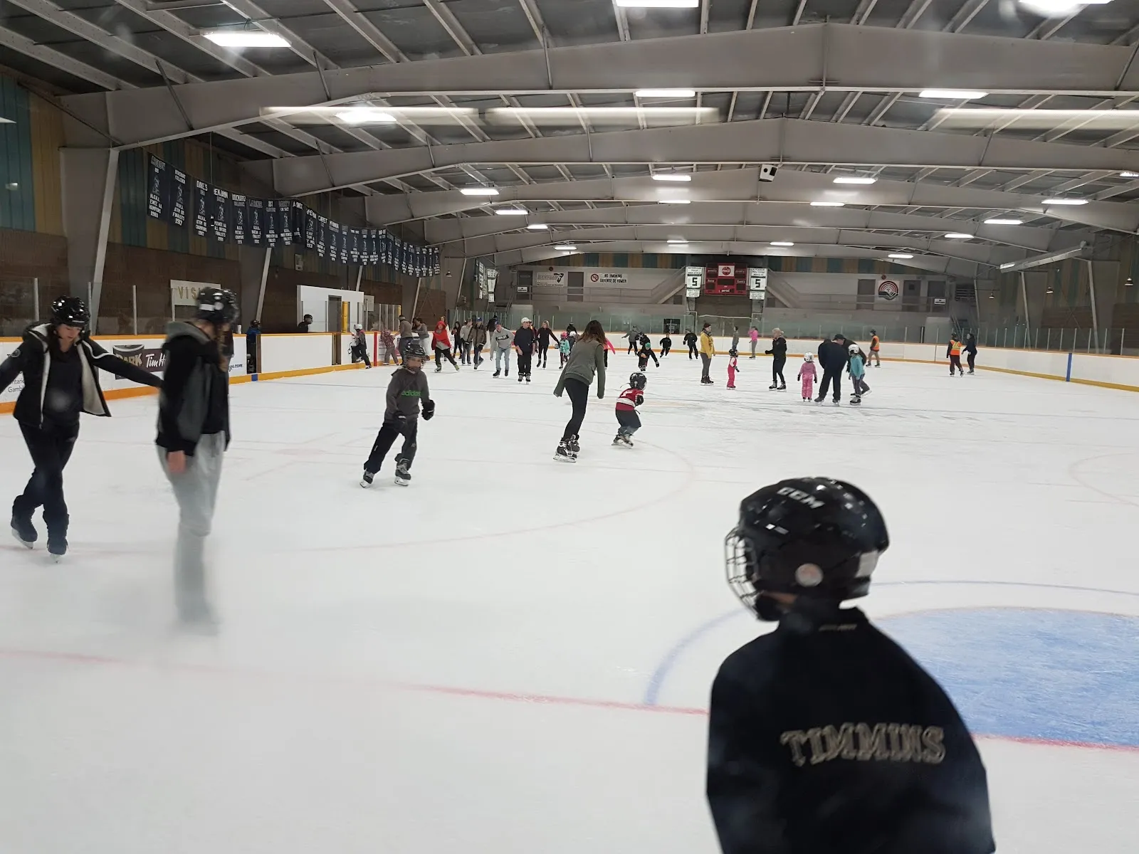 A public skating session on the ice at Mountjoy Arena: many skaters, mostly children, spread across the ice under the arched roof with banners along the boards. — Mountjoy Arena
