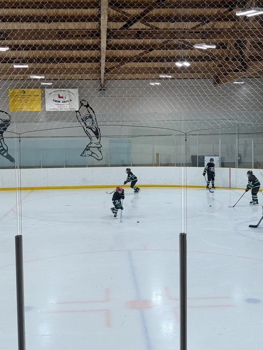 The ice at Moyer Recreation Centre seen through the glass and overhead netting: young players in a hockey practice, the wooden roof beams, a painted goalie mural and sponsor banners along the far wall. — Moyer Recreation Centre