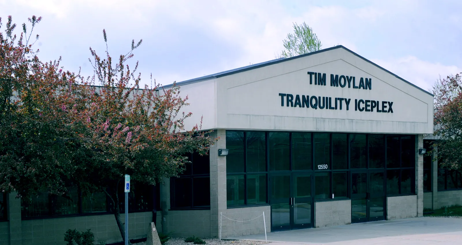 Entrada principal del edificio, un edificio bajo color arena con un frontón puntiagudo que dice "TIM MOYLAN TRANQUILITY ICEPLEX" sobre puertas de vidrio con el número 12550, y árboles en flor al frente — Moylan Ice Plex