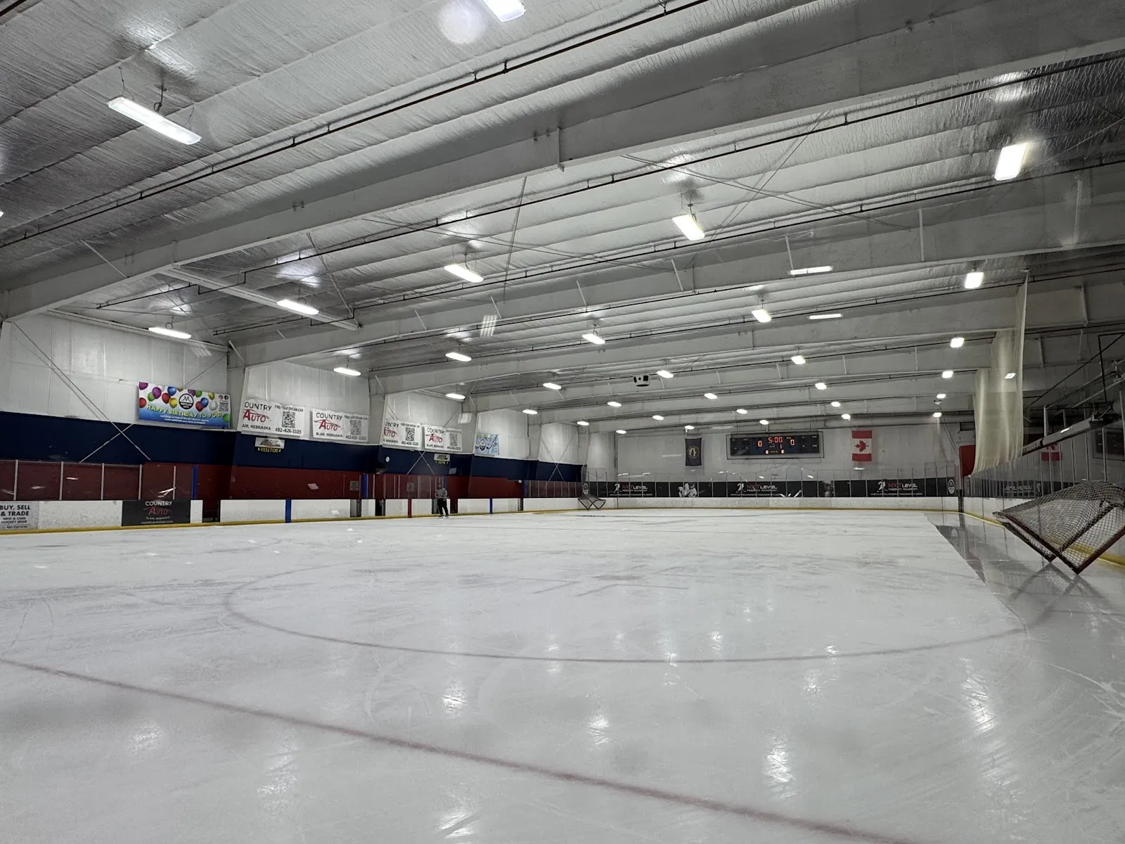 Pista vacía de tamaño NHL desde el nivel de la pista a lo largo hacia una portería de hockey y un marcador en 0-0, 5:00; vallas rojas y azules con anuncios de Country Auto (Blair, Nebraska) en las paredes y una persona caminando cerca de las vallas del fondo — Moylan Ice Plex