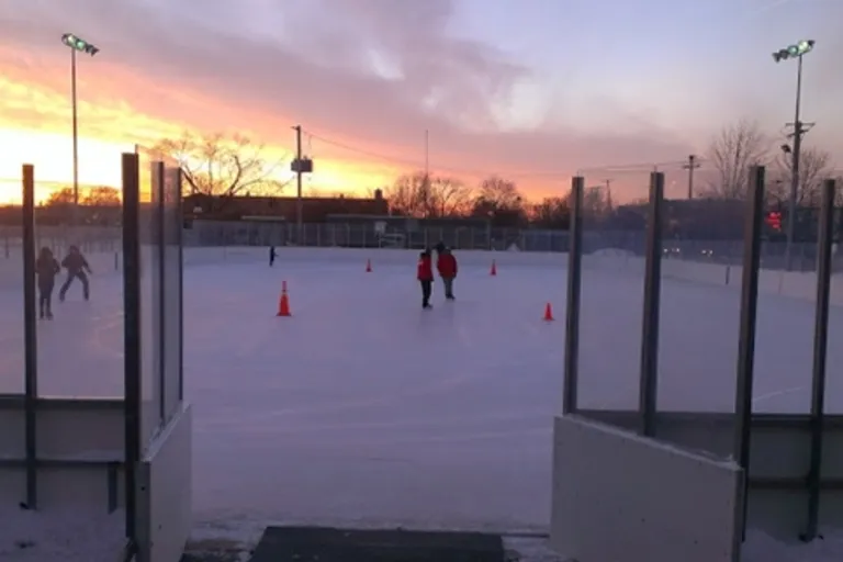 The Mount Greenwood Park ice rink at sunset seen through the boards, with a few skaters in the distance and orange cones on the ice. — Mt. Greenwood Ice Rink