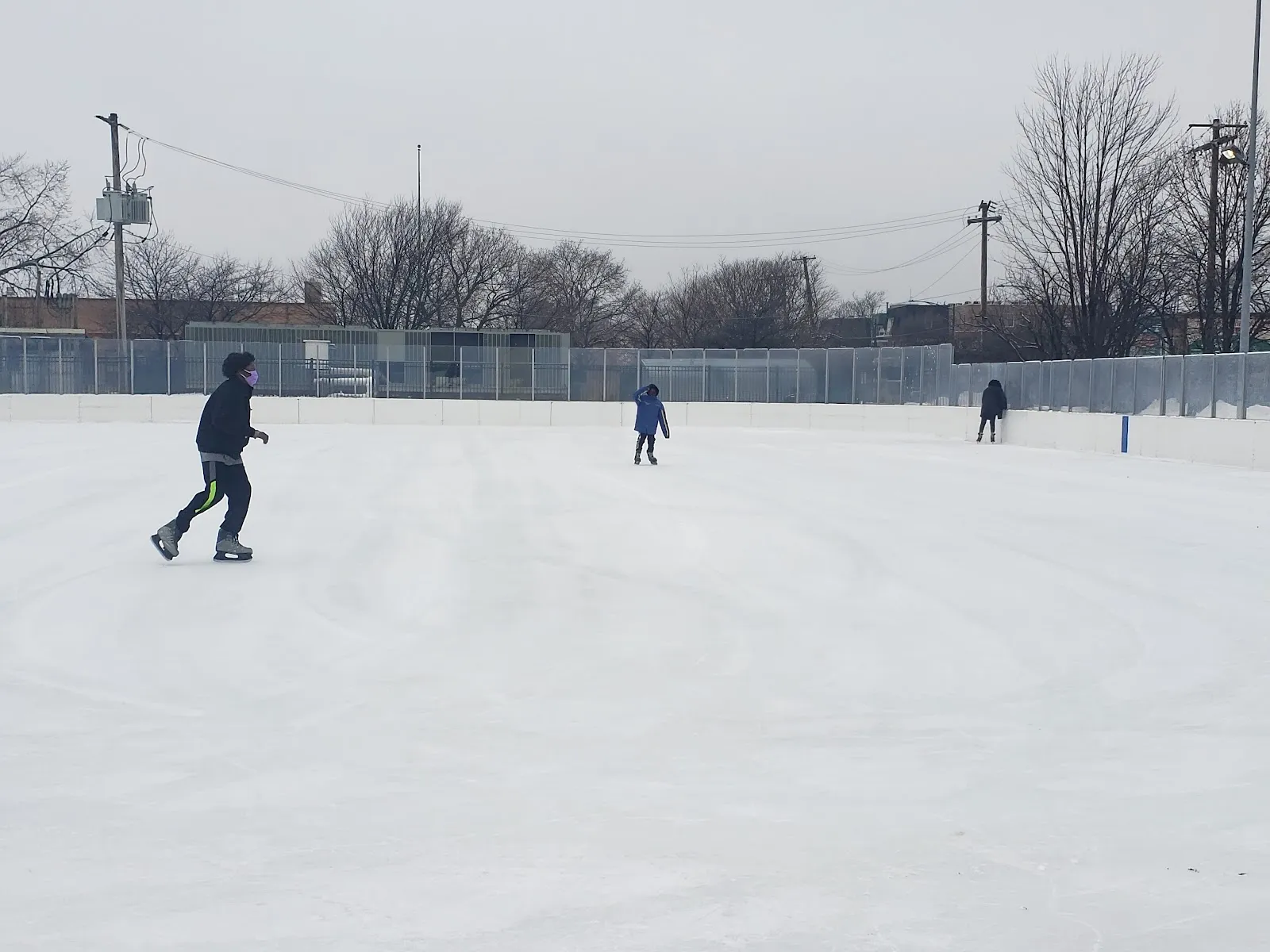 The outdoor ice surface in daylight, bounded by low boards and a chain-link fence with bare trees behind and a few skaters in the distance. — Mt. Greenwood Ice Rink