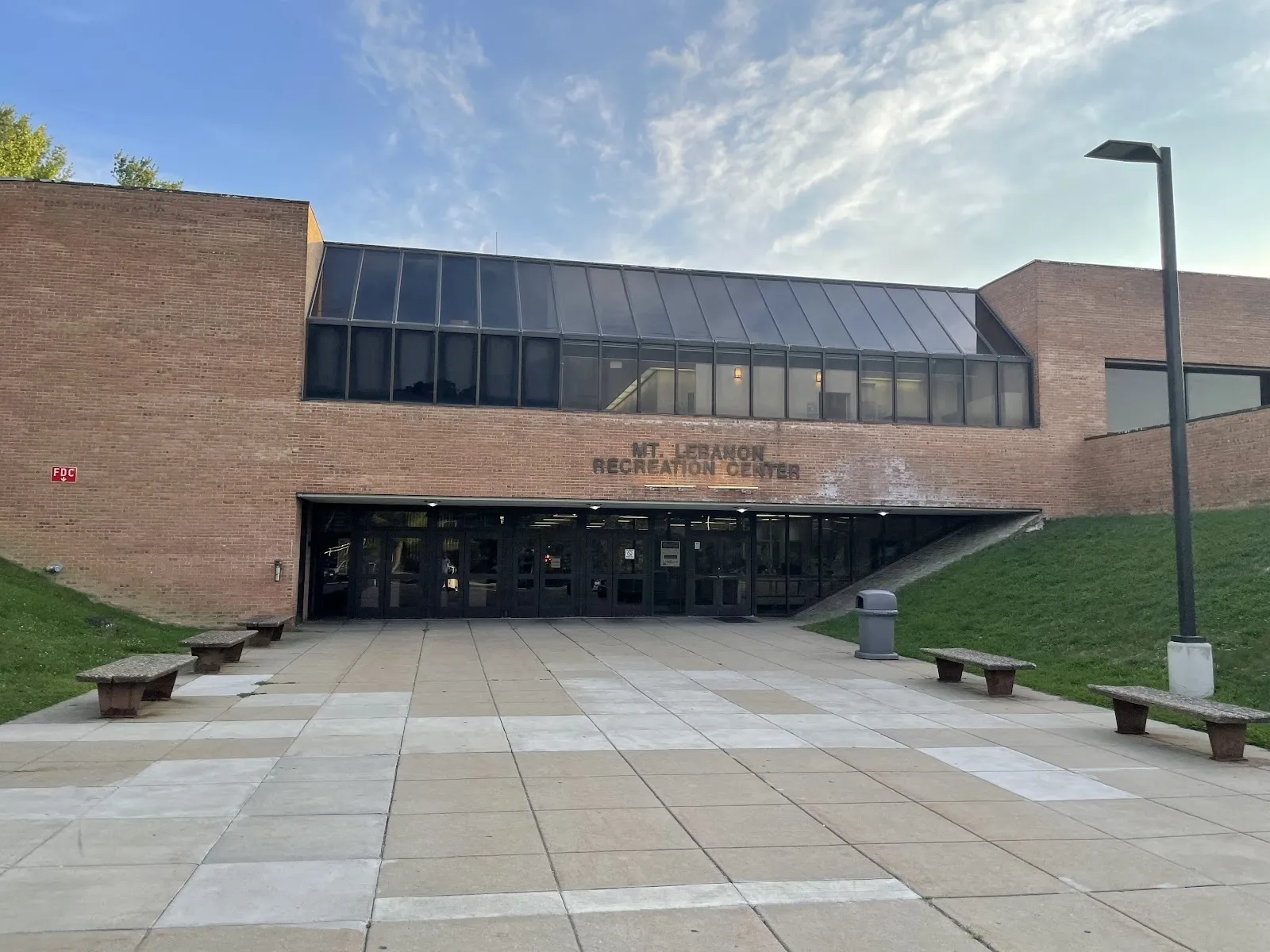 The brick front of the Mt. Lebanon recreation building with a long dark glazed band over the entrance and a paved forecourt with benches. — Mt. Lebanon Recreation Center (Frank Gill Ice Rink)