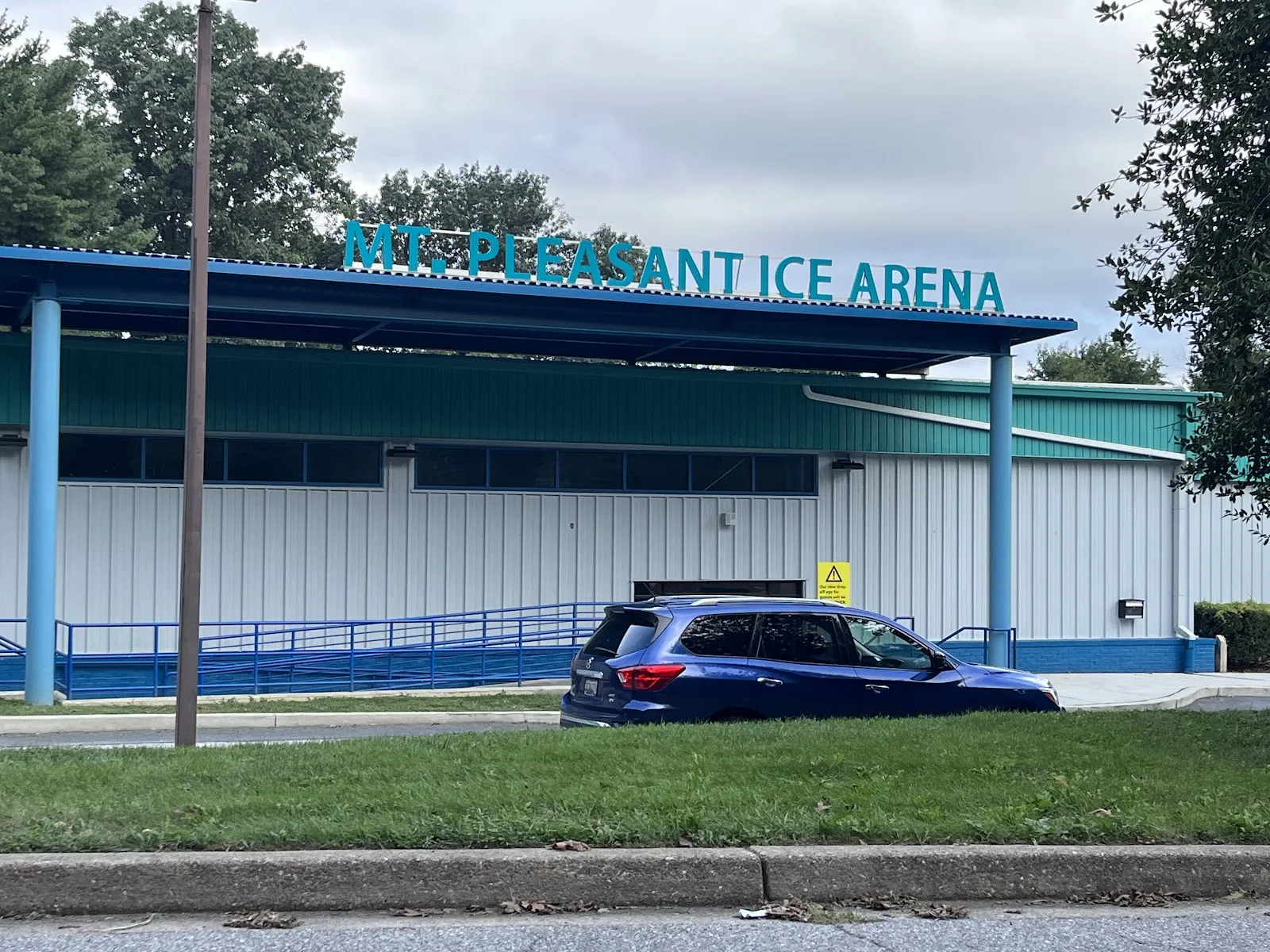 La Mt. Pleasant Ice Arena de techo azul con su nombre en letras verde azulado a lo largo de la línea del techo, vista desde la calle al otro lado del pasto — Mt. Pleasant Ice Arena