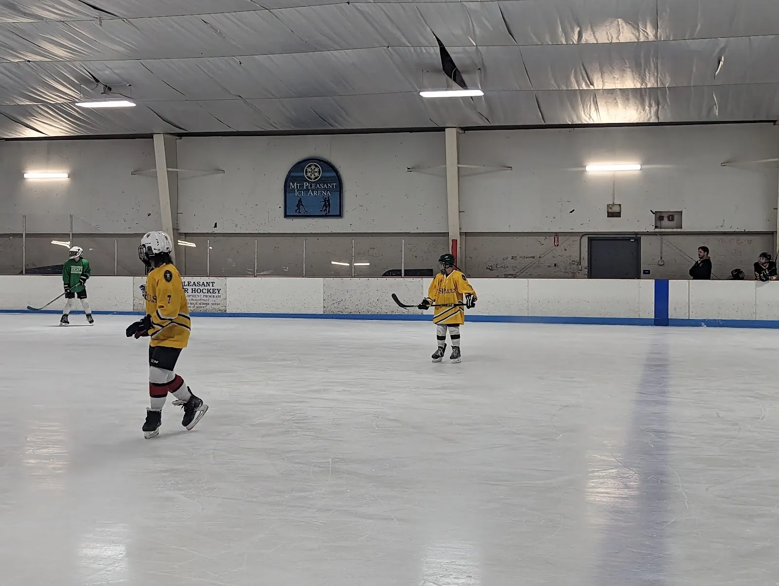 Dos jóvenes jugadores de amarillo a lo lejos sobre una pista clara y amplia, con una pared de franja azul y una placa redonda detrás de ellos bajo un techo de acero. — Mt. Pleasant Ice Arena