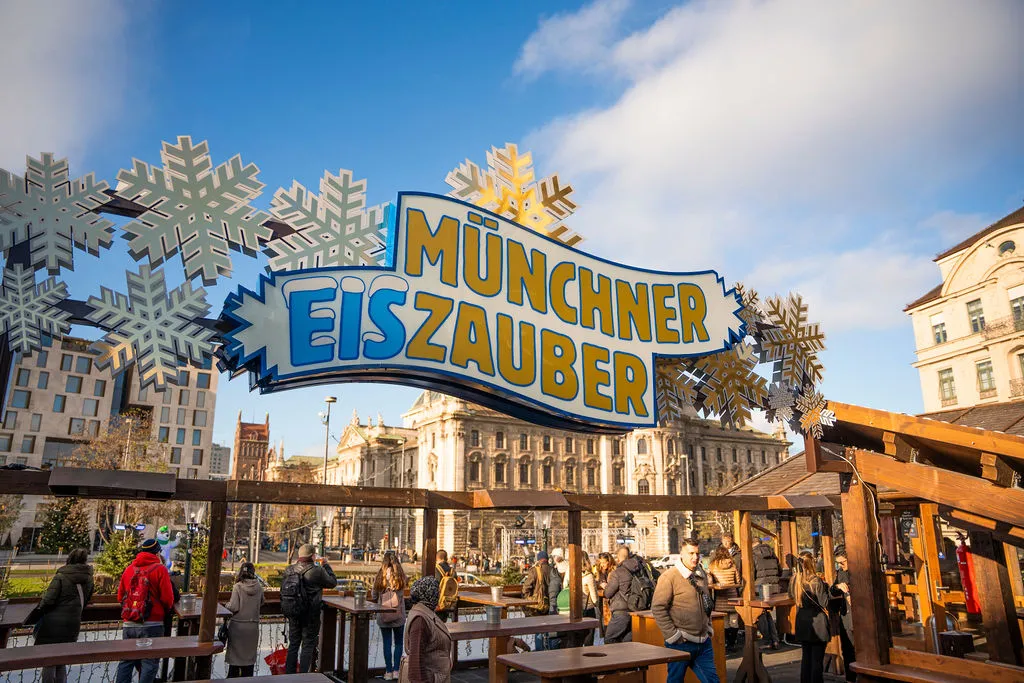 Das Münchner-Eiszauber-Schild mit Schneeflockendekoration über den hölzernen Bars und Imbisshütten am Karlsplatz (Stachus), dahinter der Justizpalast unter blauem Himmel. – Münchner Eiszauber am Stachus