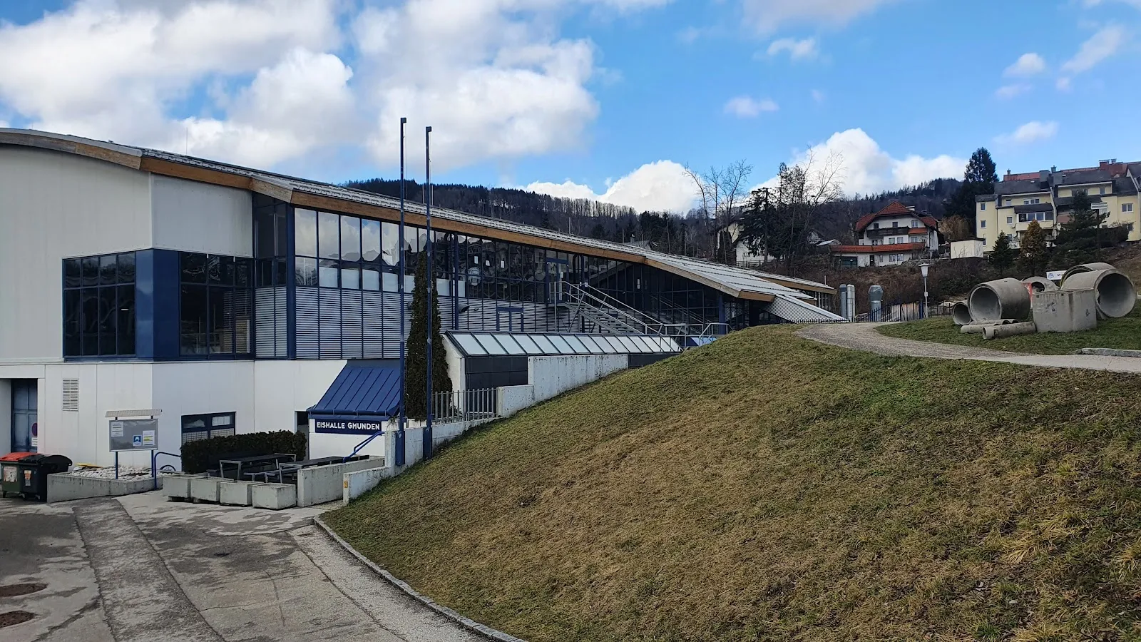 The ice hall from its hillside side: a long white building with blue trim and a pitched roof, grass embankment in front and a path up to the entrance. — Muki-Eisarena Gmunden