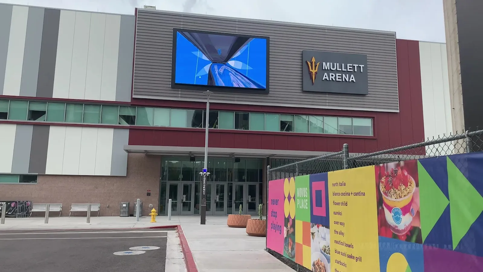 The Mullett Arena entrance at night, a wide glass-and-metal front with large video screens above the doors and a few people at the plaza. — Mullett Arena