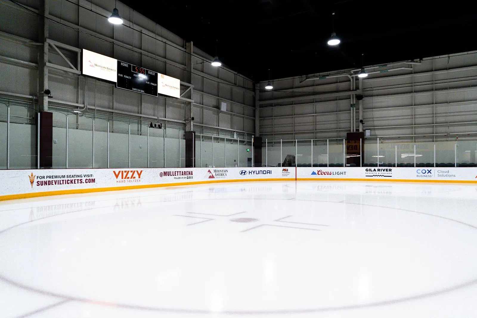 The empty Community Iceplex sheet with the center-ice logo, yellow-topped boards with sponsor panels and a scoreboard above the far end. — Mullett Arena