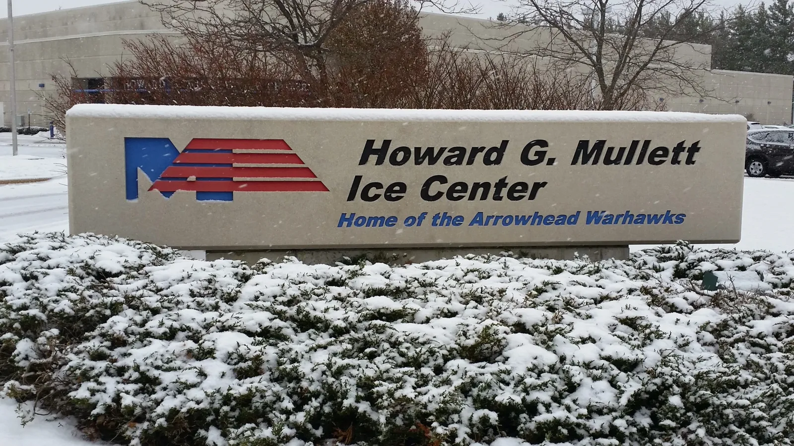 Letrero monumental que dice "Howard G. Mullett Ice Center, Home of the Arrowhead Warhawks" frente al muro exterior color arena del edificio, con nieve en el suelo — Mullett Ice Center