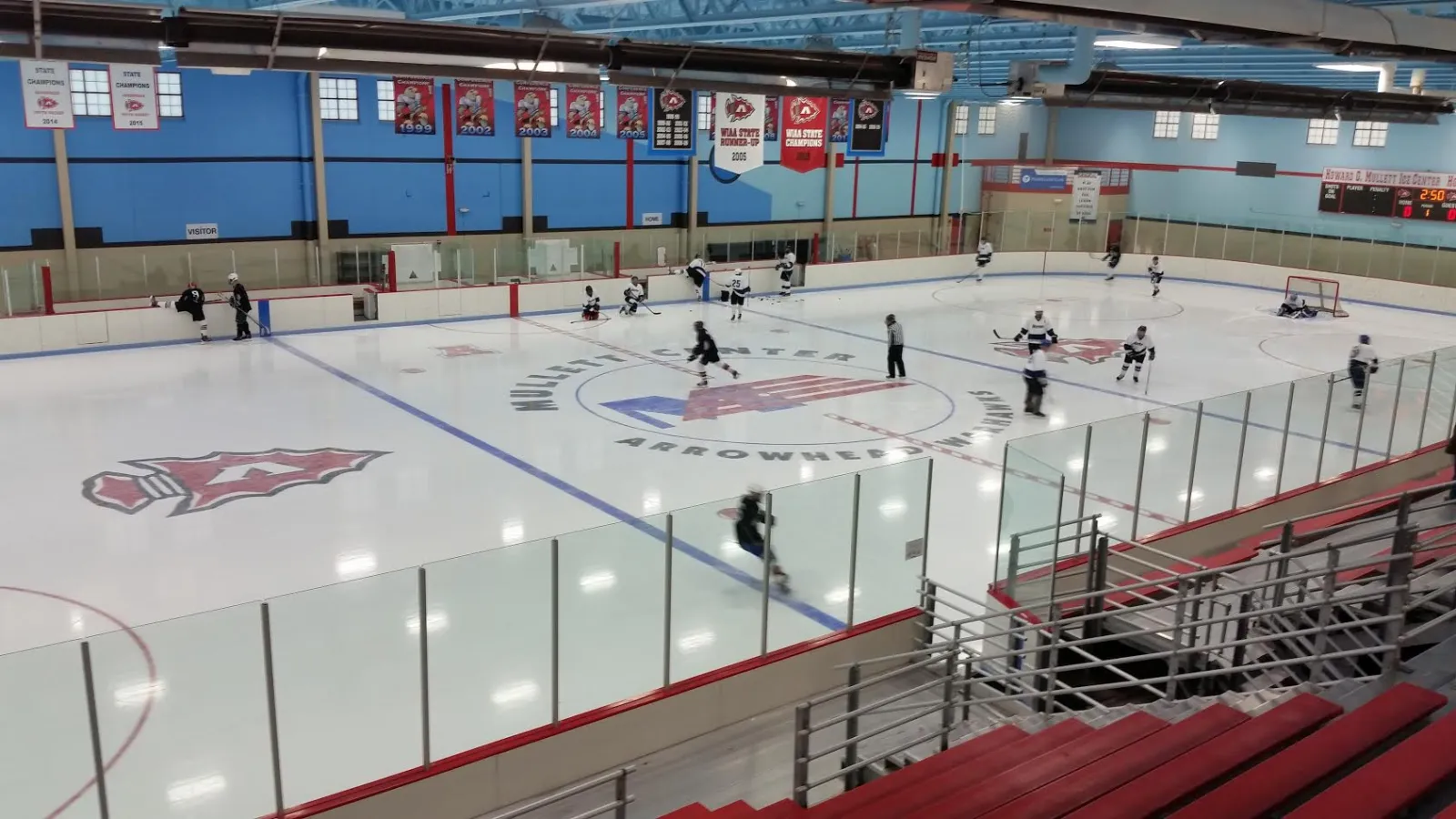 Toda la pista desde las gradas, con el logotipo de Mullett Center / Arrowhead Warhawks en el centro de la pista, mantas de campeonato en la pared azul y jugadores en el hielo durante un partido — Mullett Ice Center