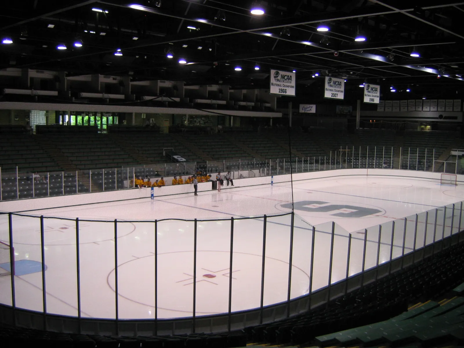 The ice surface from the seating bowl, with the Spartan S at centre ice, the boards and glass, the green seats and NCAA banners hanging from the roof. — Munn Ice Arena