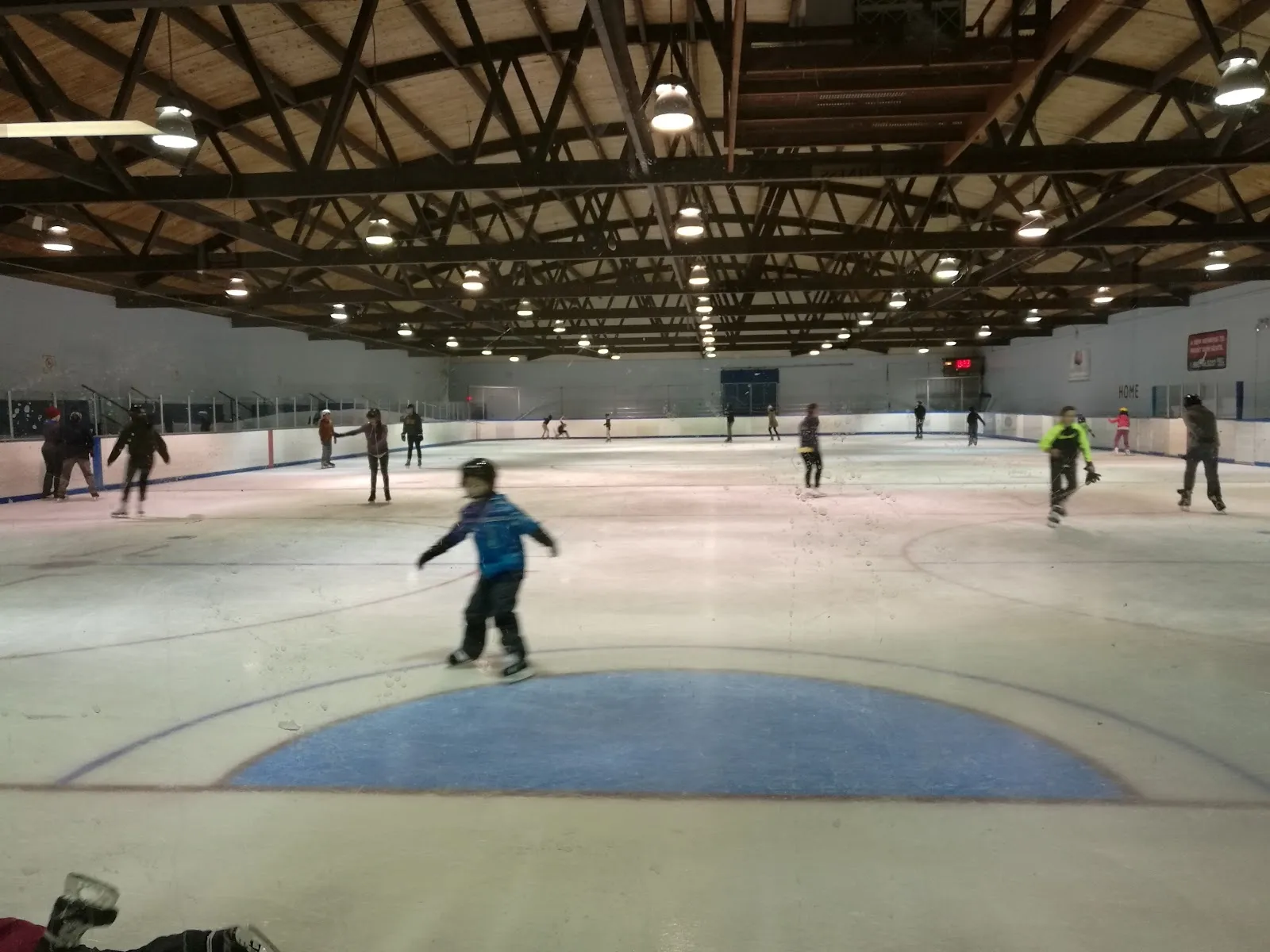 A public session on the ice under a timber-framed roof, a child skating past the blue centre circle and others spread along the far boards. — Murray Balfour Arena