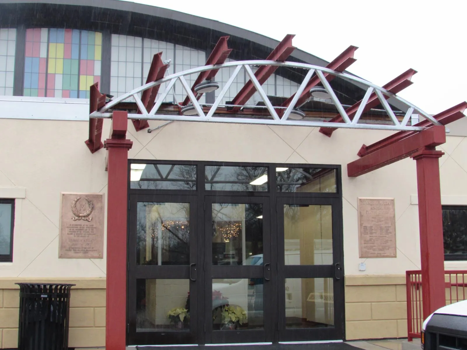 Entrada principal de Edward J. Murray Memorial Skating Center, con una marquesina de acero rojo sobre las puertas de vidrio y una placa conmemorativa de bronce y una placa de dedicatoria a los lados de la entrada; arriba se ven la línea en arco del techo de la pista y una ventana alta estilo vitral. — Edward J. Murray Memorial Skating Center
