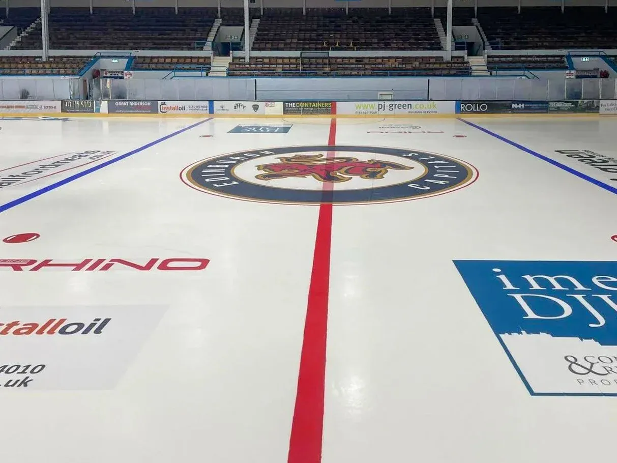 The empty ice surface of the Murrayfield arena, with its centre-ice crest, red centre line, blue lines and the boards and sponsor ads; no people. — Murrayfield Ice Arena