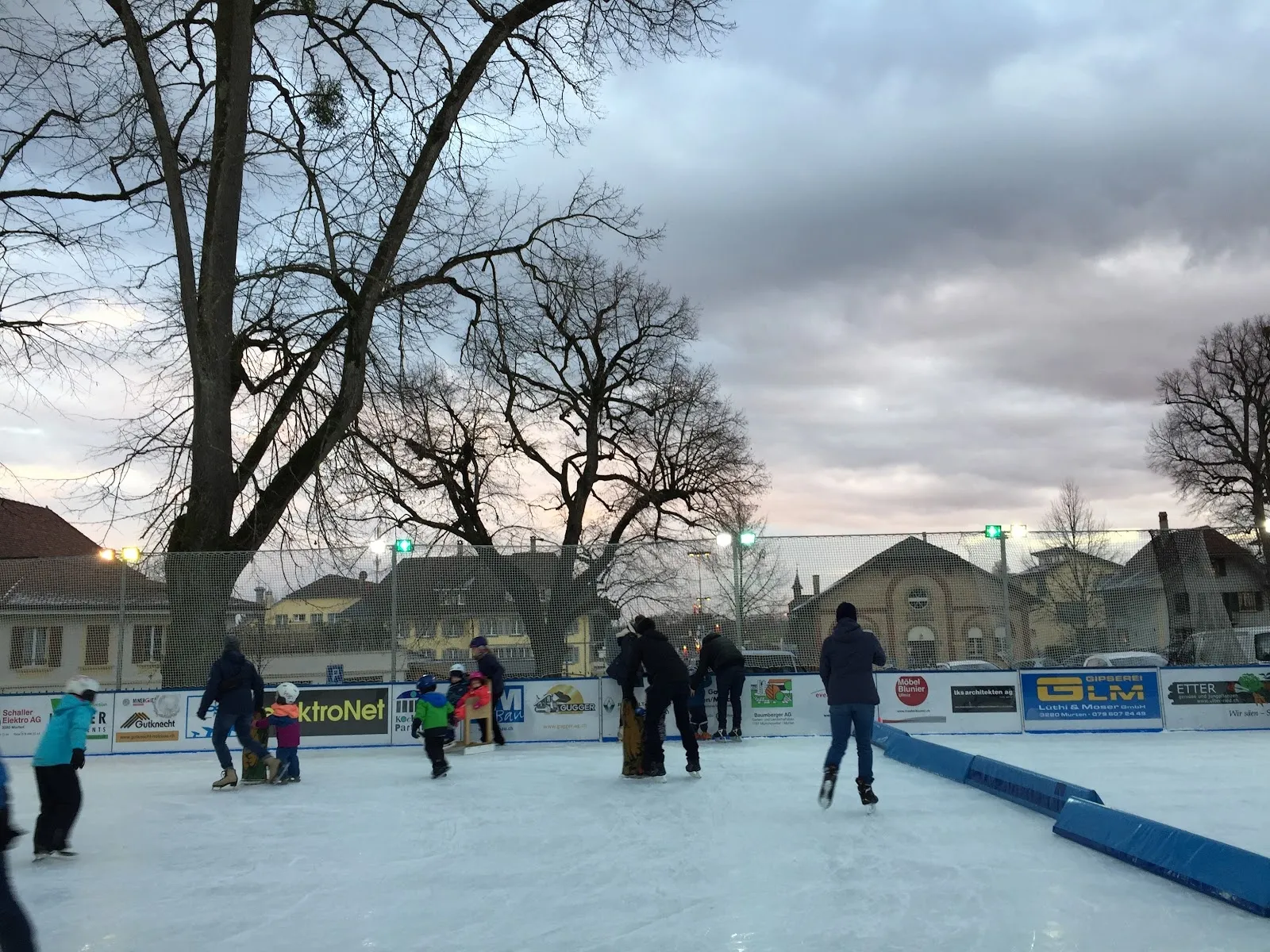 The Murten on Ice rink at the Berntorplatz with skaters on the ice and the town's houses and bare trees behind, under an overcast sky. — Murten on Ice