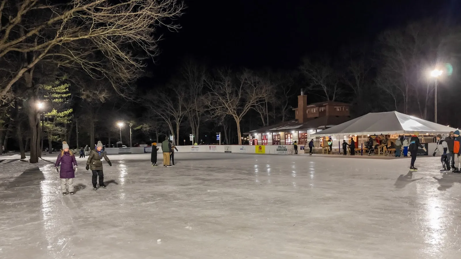 Skaters on the lit outdoor rink at night with the lodge and a tent behind — Muskegon Luge Adventure Sports Park Ice Rink