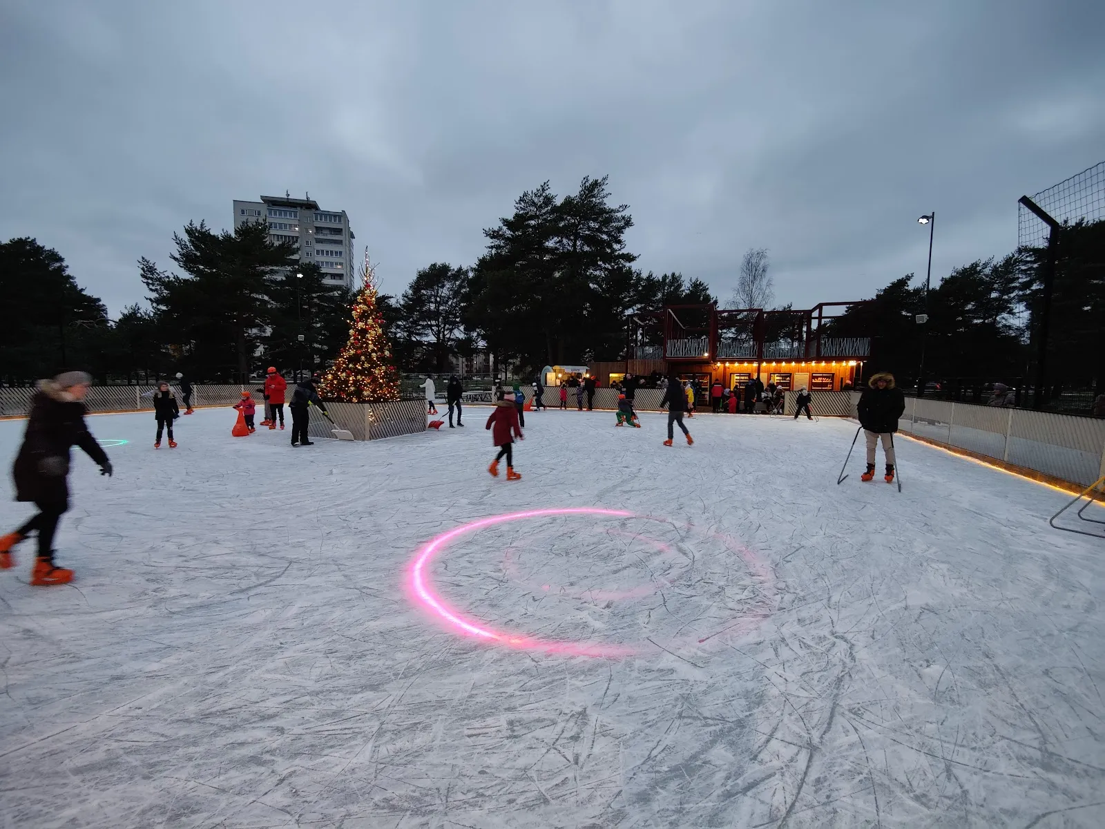 The Mustamäe outdoor ice at dusk with a few skaters, a lit Christmas tree and the lights along the rink edge. — Mustamäe uisupark