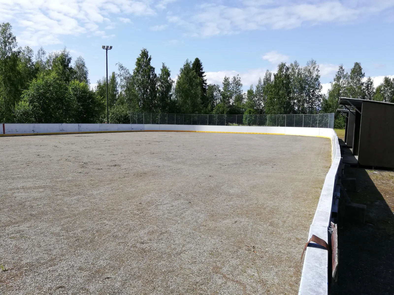 The boarded Muuruvesi rink in summer, with a light mast and trees behind the boards. — Muuruveden jääkiekkokaukalo