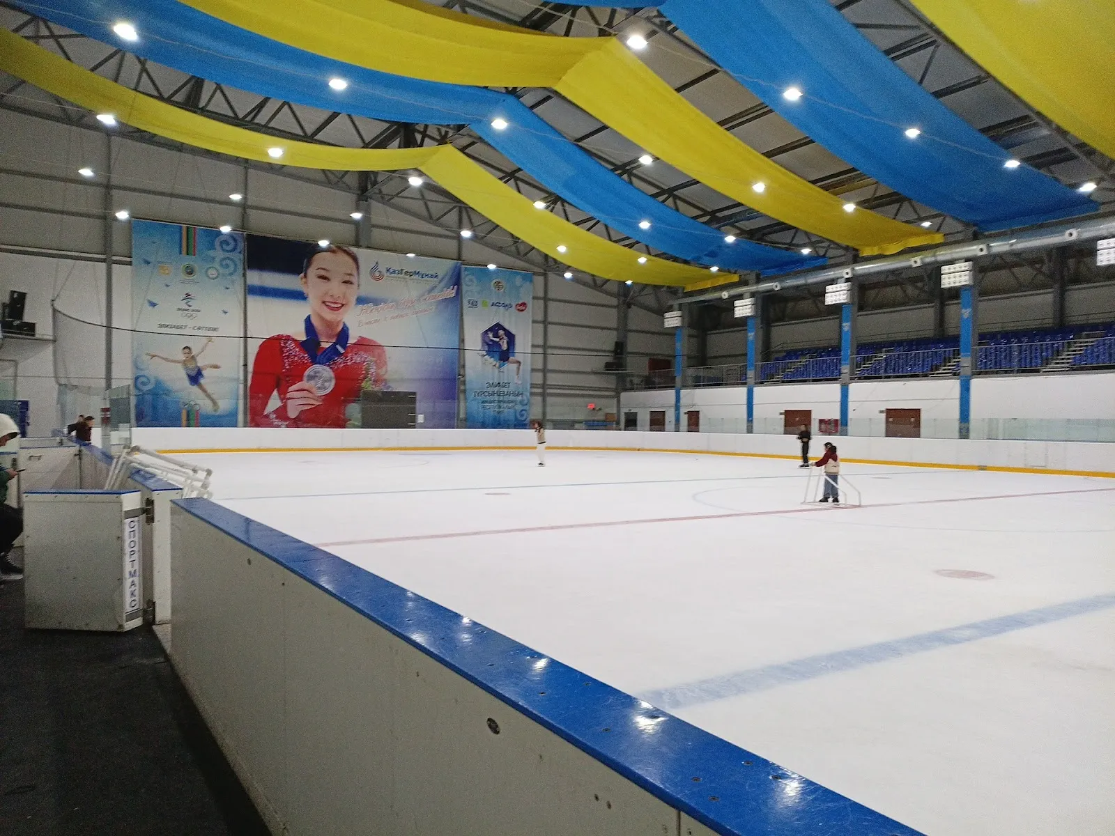 The ice sheet of the Muz Aidyny rink in Kyzylorda under its yellow and blue ceiling drapes, with a figure skater banner on the wall — Muz Aidyny Ice Rink