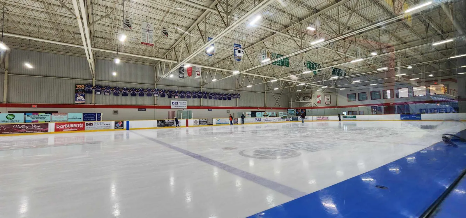 El hielo de Deslaurier Arena desde las vallas de la esquina, con mantas colgando de las armaduras de acero del techo y anuncios de patrocinadores a lo largo del lado opuesto. — myFM Centre (Renfrew)