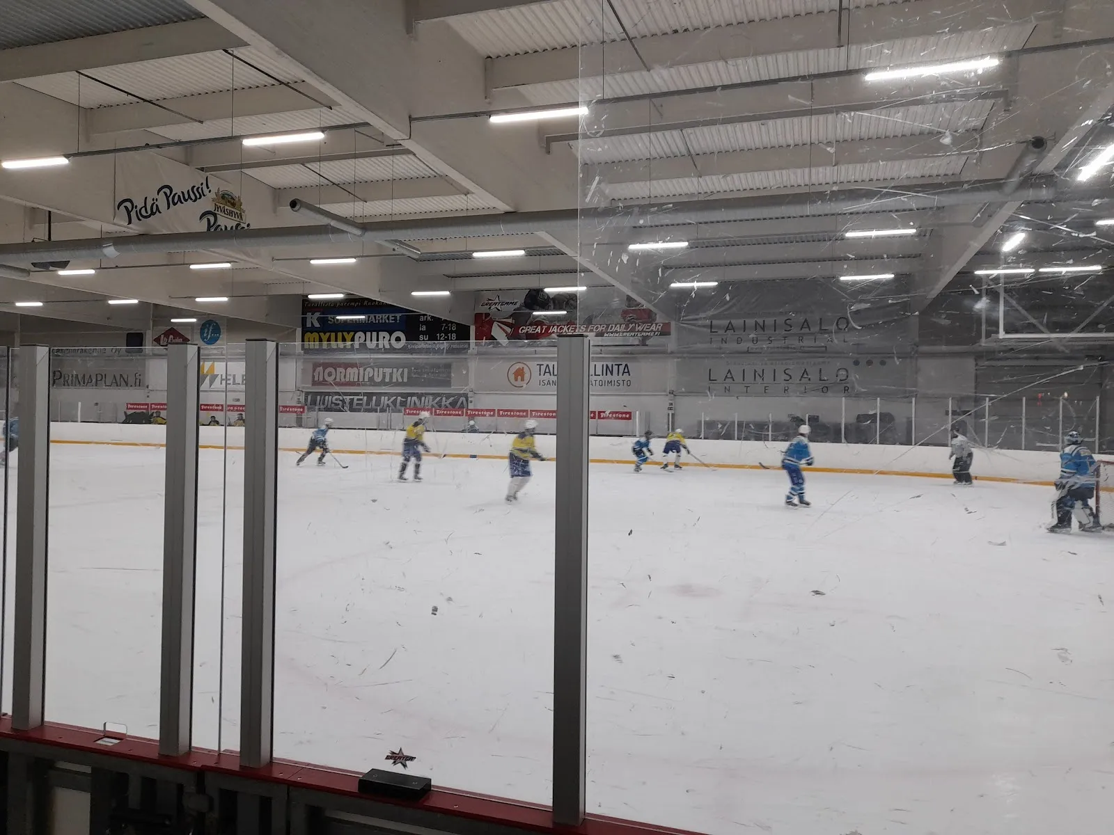 Spelare på isen i Myllypuron jäähalli i Helsingfors, sedda över hela isytan bakom plexiglaset och sargen under halltaket. – Myllypuron jäähalli