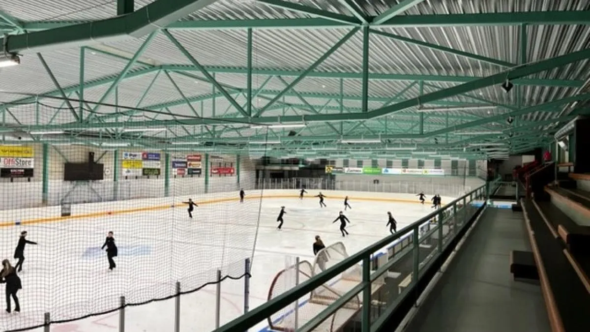 The ice of Myyrmäen jäähalli 2 under its green steel-truss roof, seen from the gallery along the boards, with skaters spread across the ice — Myyrmäen jäähalli 2