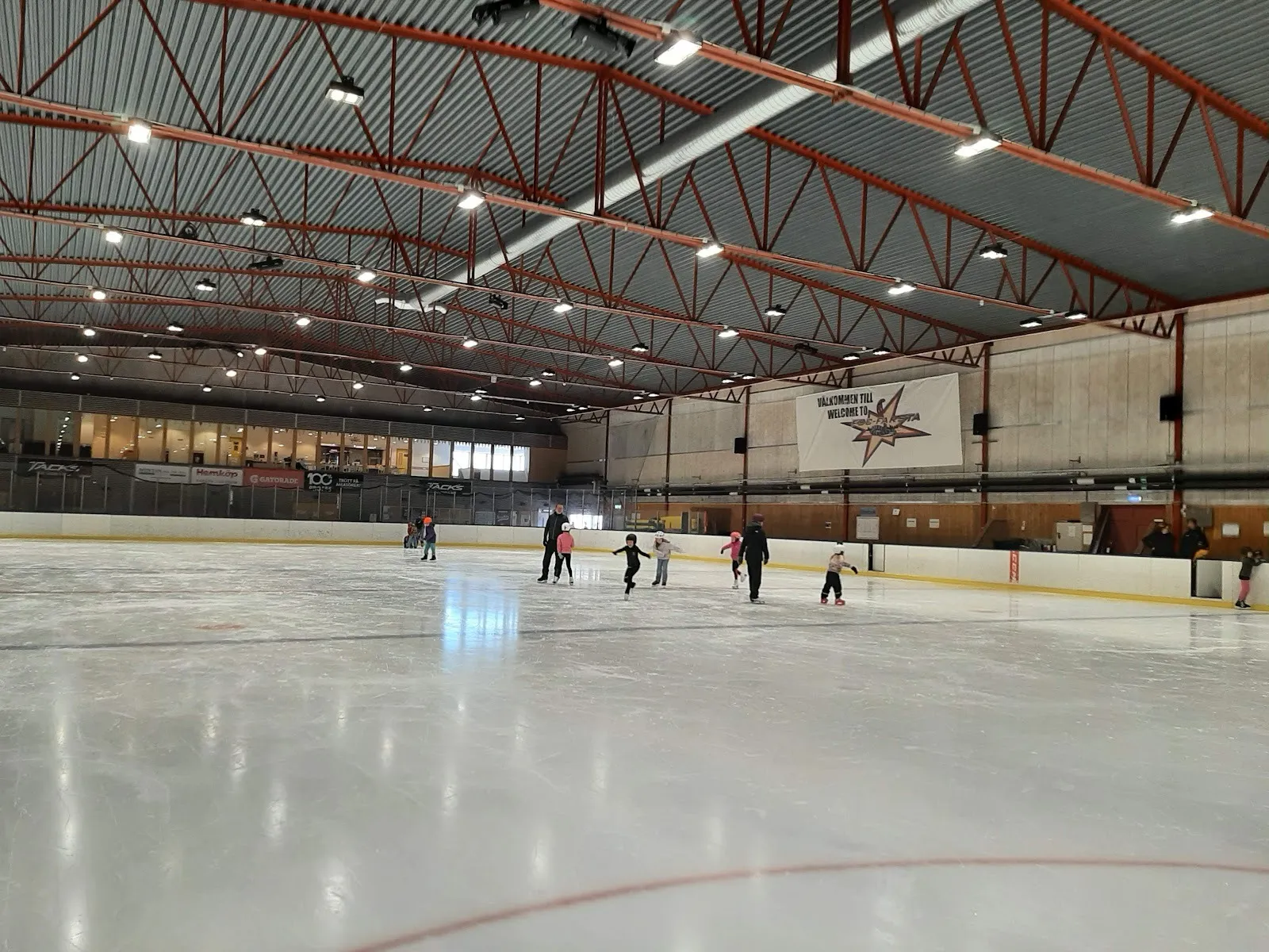 The Nacka ice hall during a session, skaters across the ice under the red steel roof trusses with the stands behind the boards. — Nacka ishall